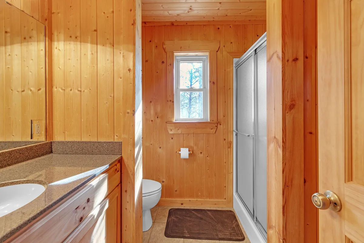 This bathroom features wooden paneling, a vanity with a sink, a shower stall, and a window that lets in natural light. The space feels fresh and functional for morning routines or winding down after a busy day.