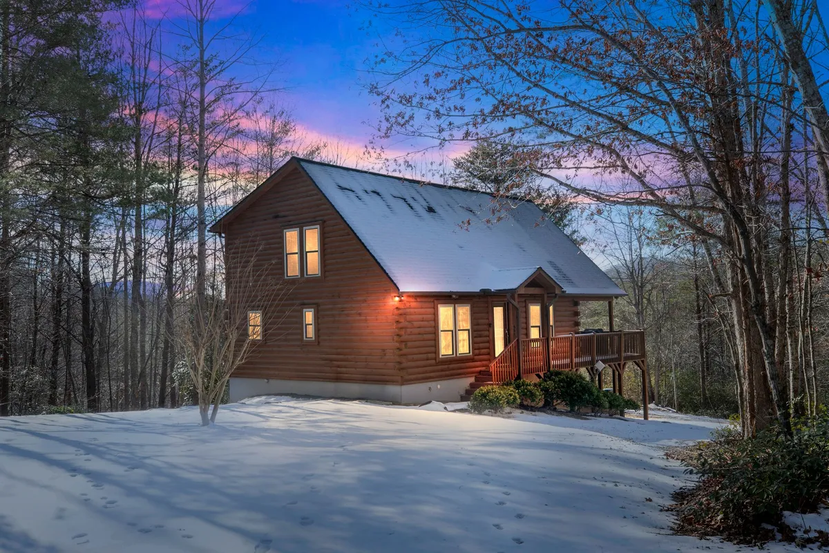 A warmly lit wooden cabin stands amidst a snow-covered landscape, emitting a cozy glow. The scene is peaceful, with snow play visible outside, and the setting sun or early evening sky adding to the ambiance.