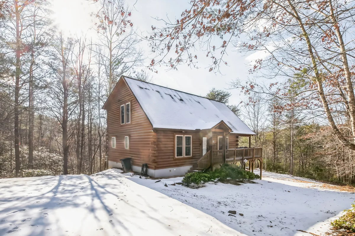 A serene shot of the log cabin's exterior, featuring snowy ground and mature trees. The image implies opportunities for winter walks, outdoor gatherings, and enjoying the landscape from the porch, reinforcing the spirit of group togetherness and activities.