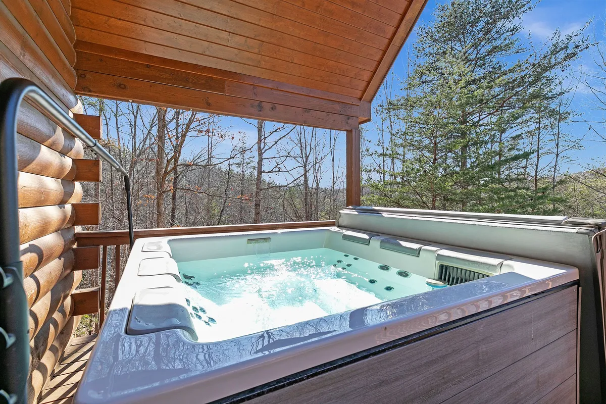 A tranquil outdoor hot tub nestled among trees, surrounded by a wooden structure that suggests privacy and connection to nature. The setting invites relaxation and socializing under the open sky, with the deck seamlessly blending indoor comfort with outdoor serenity.