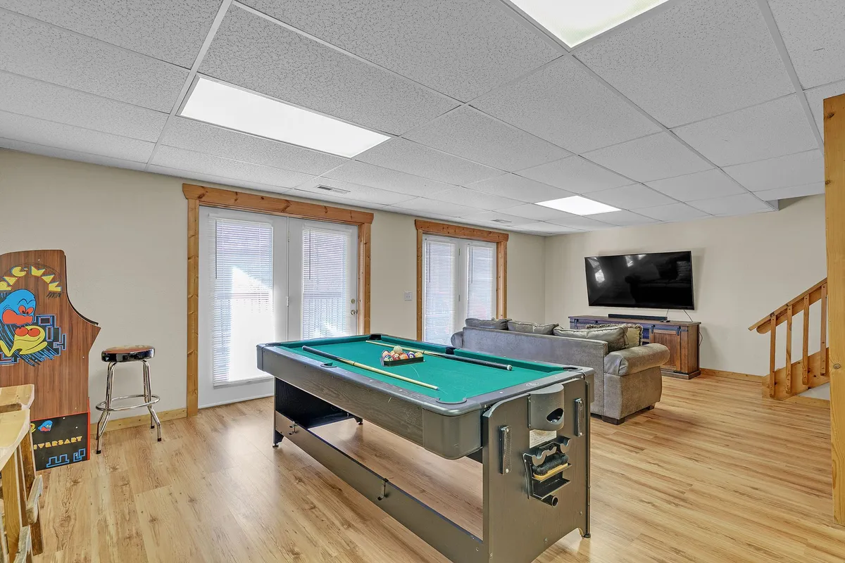 A recreational basement scene featuring a pool table, spacious sofa, large TV, and sliding doors, set in a modern, wood-paneled environment. The area is clearly set up for both gaming and lounging, with an open, social atmosphere.