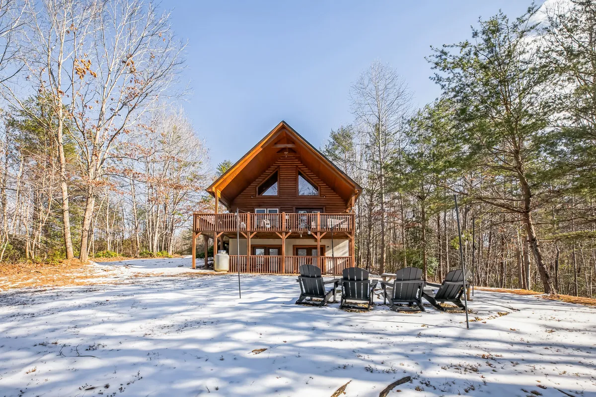 The image showcases a classic log cabin embraced by tall trees, with a spacious deck and a fire pit surrounded by Adirondack chairs. Fresh snow blankets the ground, evoking a serene winter retreat. The overall scene is cozy and inviting, highlighting outdoor gathering spaces perfect for relaxation and enjoying nature.