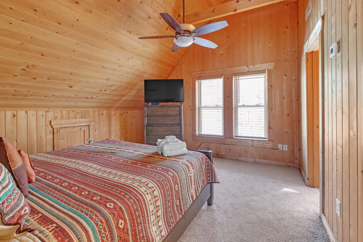 A welcoming bedroom with a colorful bed, wooden walls, and ceiling. Sunlight streams through large windows, flooding the room with natural light and offering glimpses of the outdoors, perfect for relaxed mornings together.