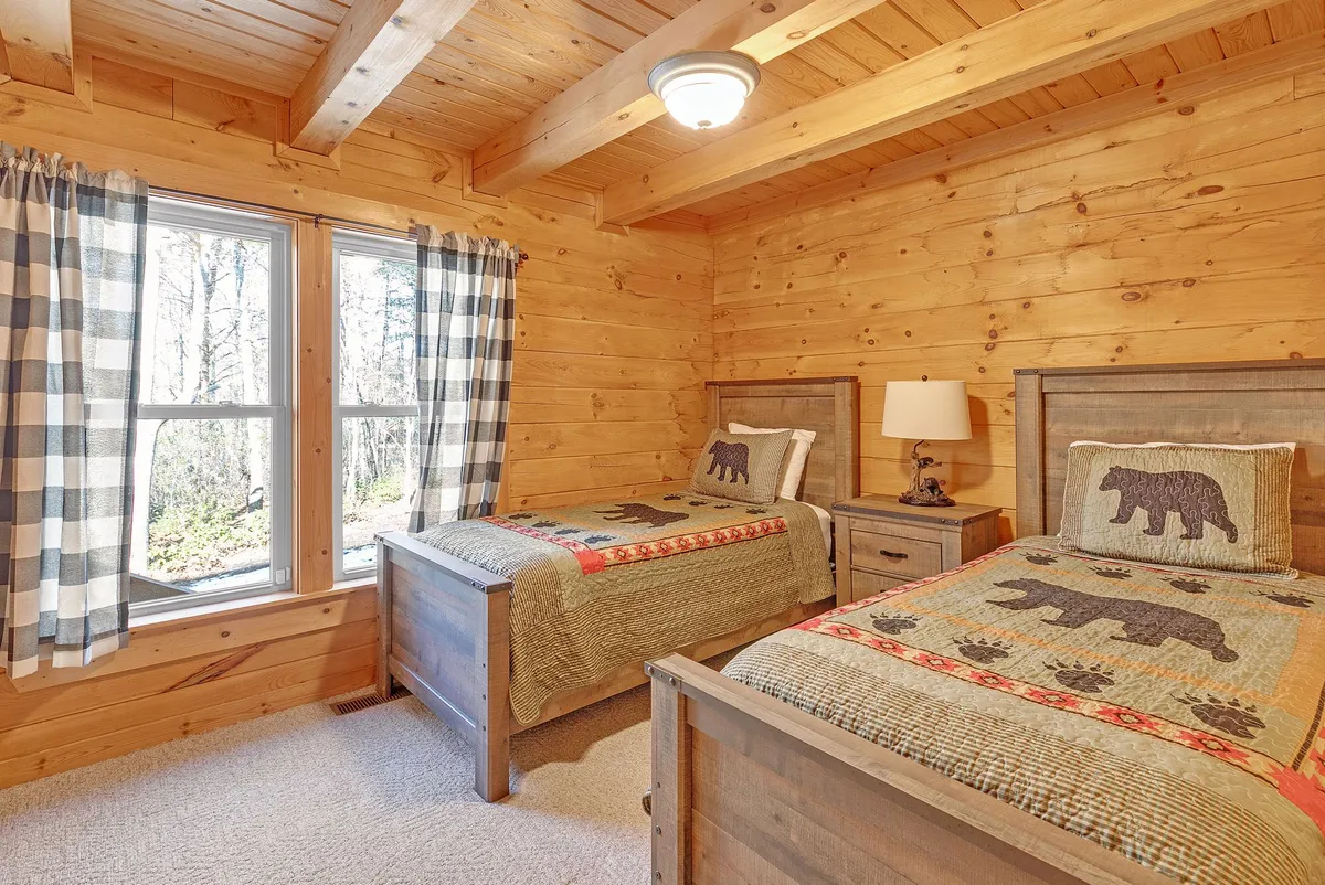 The image features a bedroom with wooden beds topped with bear-themed quilts, large windows allowing natural light to fill the room, and checkered curtains. The setting is playful yet restful—ideal for families or groups seeking comfort and a touch of whimsical decor.