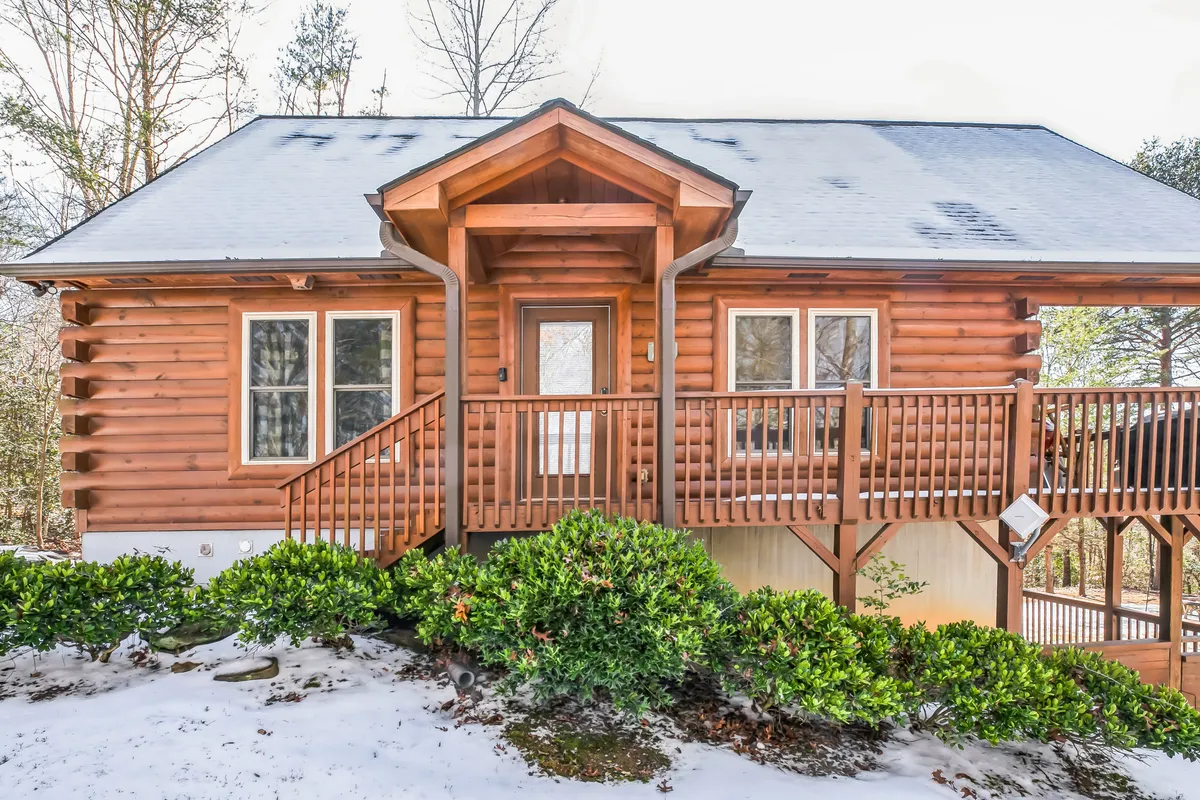 The log cabin's exterior with a wooden deck, front door, and vibrant greenery surrounding the structure, suggesting a welcoming retreat nestled in nature. The open deck is ideal for grilling, relaxing with family, or prepping for a day at the nearby lake or park.