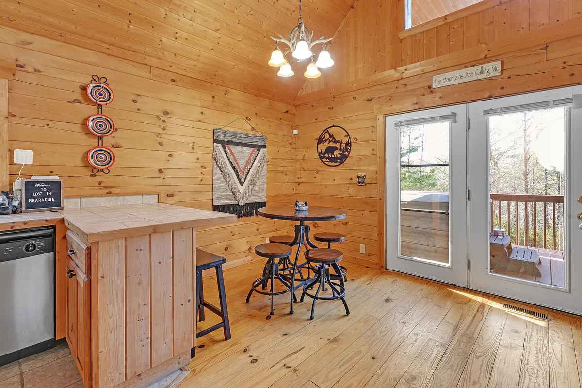 A dining area with a round table positioned near a wooden bar counter, accented by decorative wall hangings and large windows that offer views of the outdoors. The scene exudes warmth and encourages both casual meals and meaningful conversations.