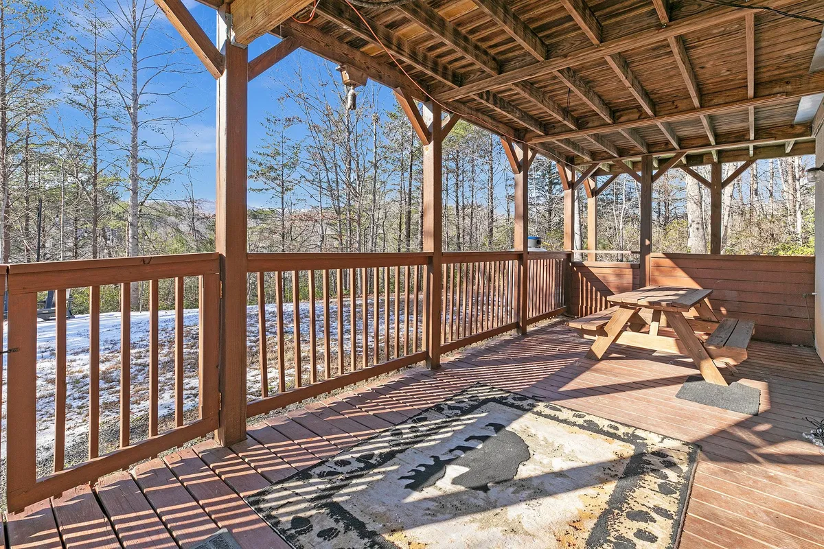 A spacious outdoor deck featuring a wooden railing, picnic table perfect for family meals or game time, a rug with a bear motif, and lush surrounding trees that evoke a sense of being immersed in nature. The setup invites groups to gather for outdoor dining, relaxation, and storytelling.