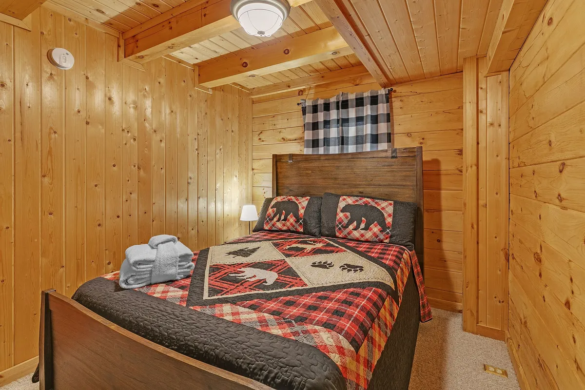 This image shows a cozy bedroom with a bed dressed in a patterned quilt, set against warm wooden walls and ceiling beams. A soft lamp glows on the nightstand, creating an inviting and restful atmosphere perfect for unwinding after an adventurous day.