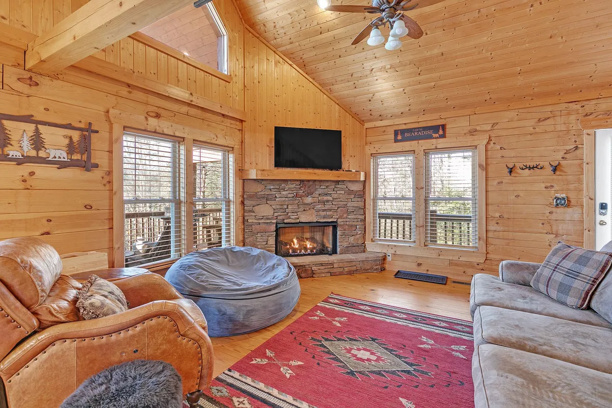 A warm, inviting basement recreation room with a plaid couch, bean bag chair, a cozy fireplace, large windows bringing in natural light, and rustic wooden beams overhead. The scene is perfect for group activities like movie nights, games, and fireside chats after a day of adventure.