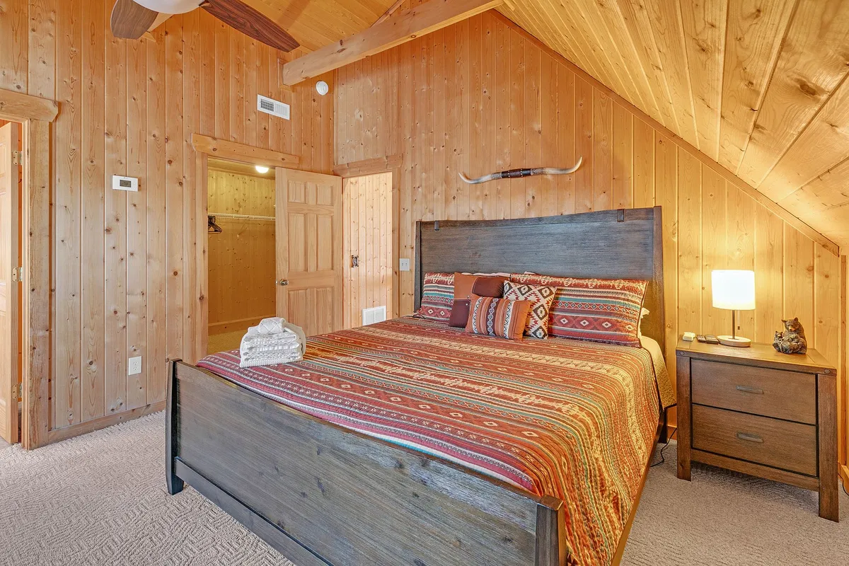 A warm, rustic bedroom featuring a wooden bed with colorful bedding, surrounded by wood paneling. A lamp glows softly on the nightstand, creating a cozy, intimate ambiance. Gentle natural light hints at mountain views beyond.